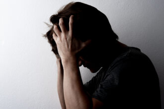 A man gripping his head in pain and frustration; most of the man is in sillhouette against a white background, only his arm really visible.