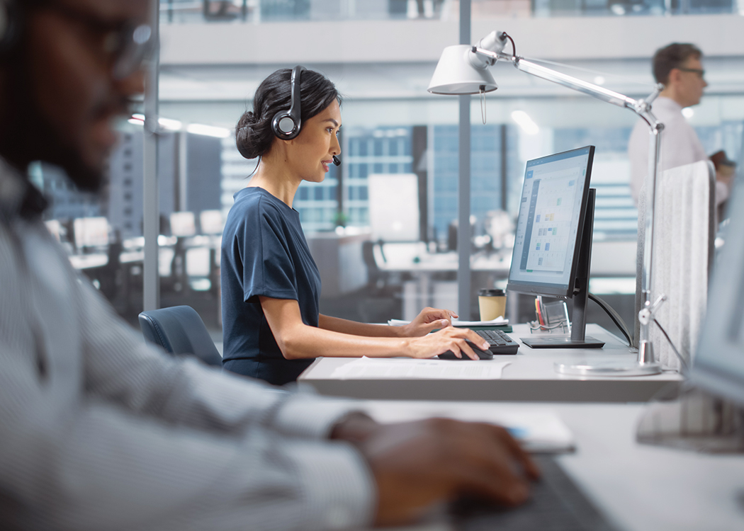 A photo of an Asian woman wearing a headset and working on a computer in an open-air office. In the bottom left corner in the foreground, there is also an out-of-focus African American man.