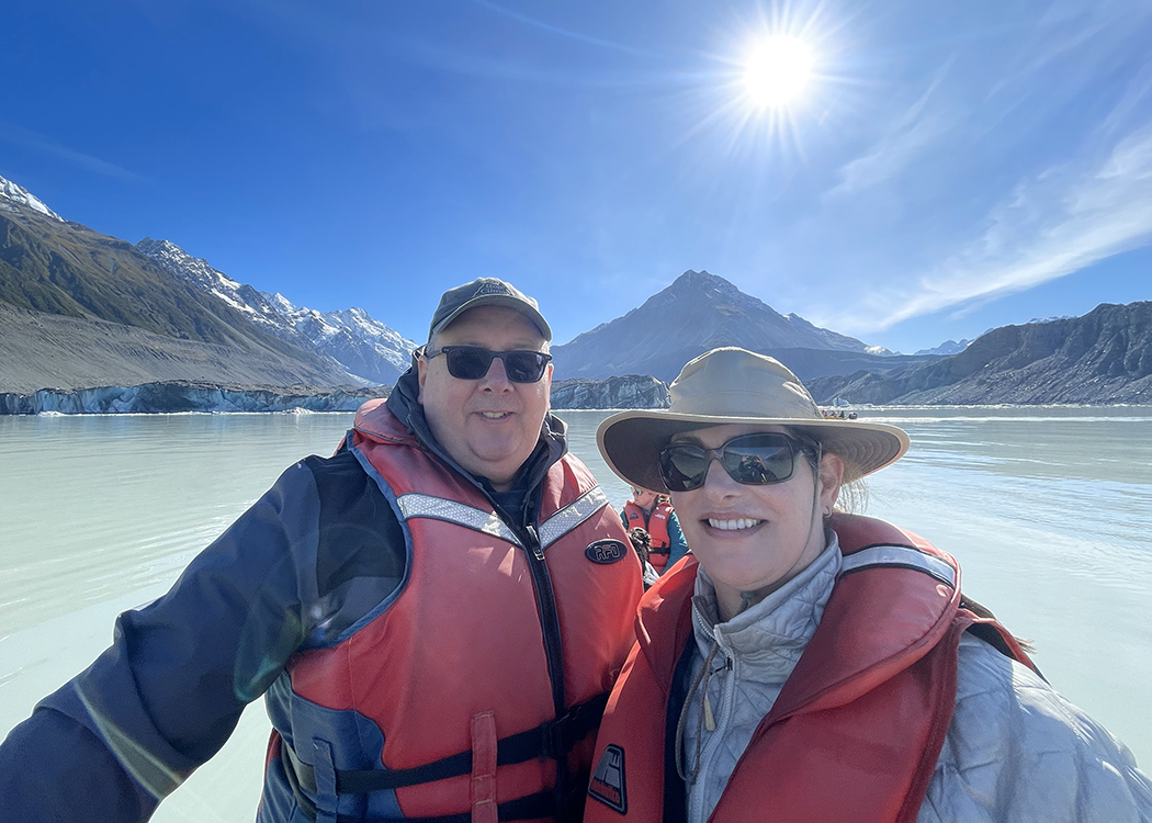 A photo of NOSSCR CEO Steve Gardner and his wife Cheryl Levine in a boat on a lake nestled between mountains. There is a bright blue sky, and the sun is visible; Steve and Cheryl are both wearing life jackets, hats, and sunglasses.