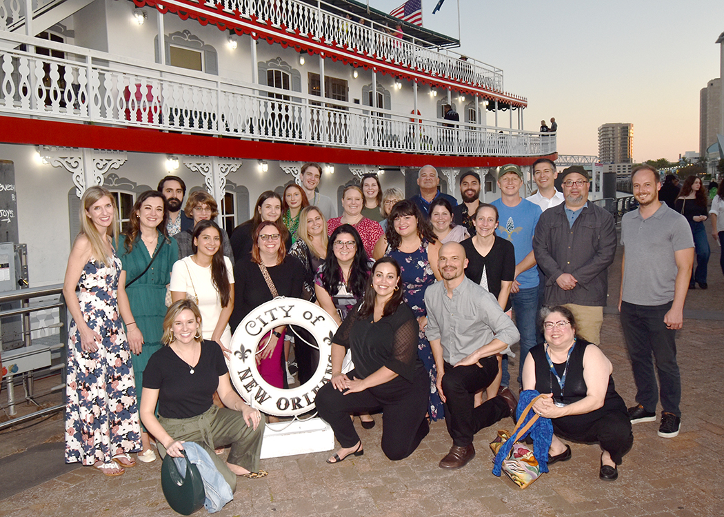 2023 NextGen attendees are posing on a dock in front of a red and white riverboat. Attendees are standing and kneeling to fit everyone in, and they are surrounding a statue in the shape of a life preserver wit the words "City of New Orleans" emblazoned on it.